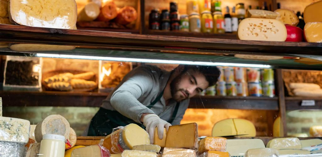 Young latin american man arranging the cheese refrigerator display at a delicatessen