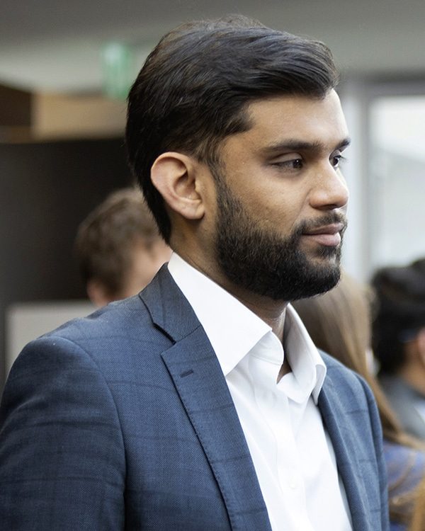 Side view of mid-30s Asian male with dark hair and beard wearing a blue suit. He is looking to the right while in conversation.